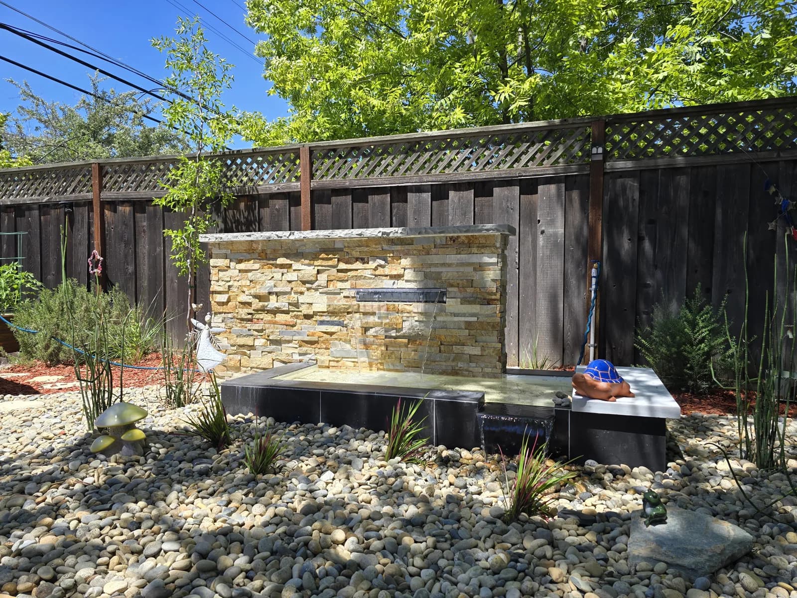 Stacked-stone wall fountain in a river rock bed in front of a wood fence