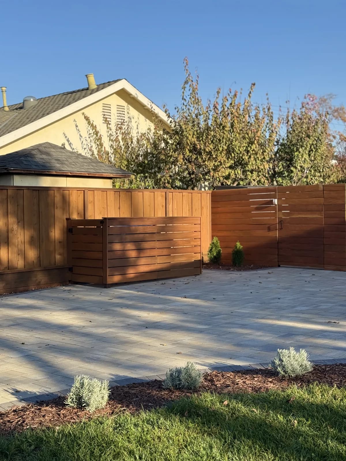 Horizontal redwood fence and gate beside a new paver patio with young plantings