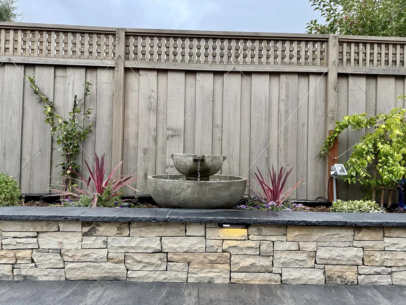 Tiered bowl fountain on a stone-veneer seat wall against a cedar fence with lattice top