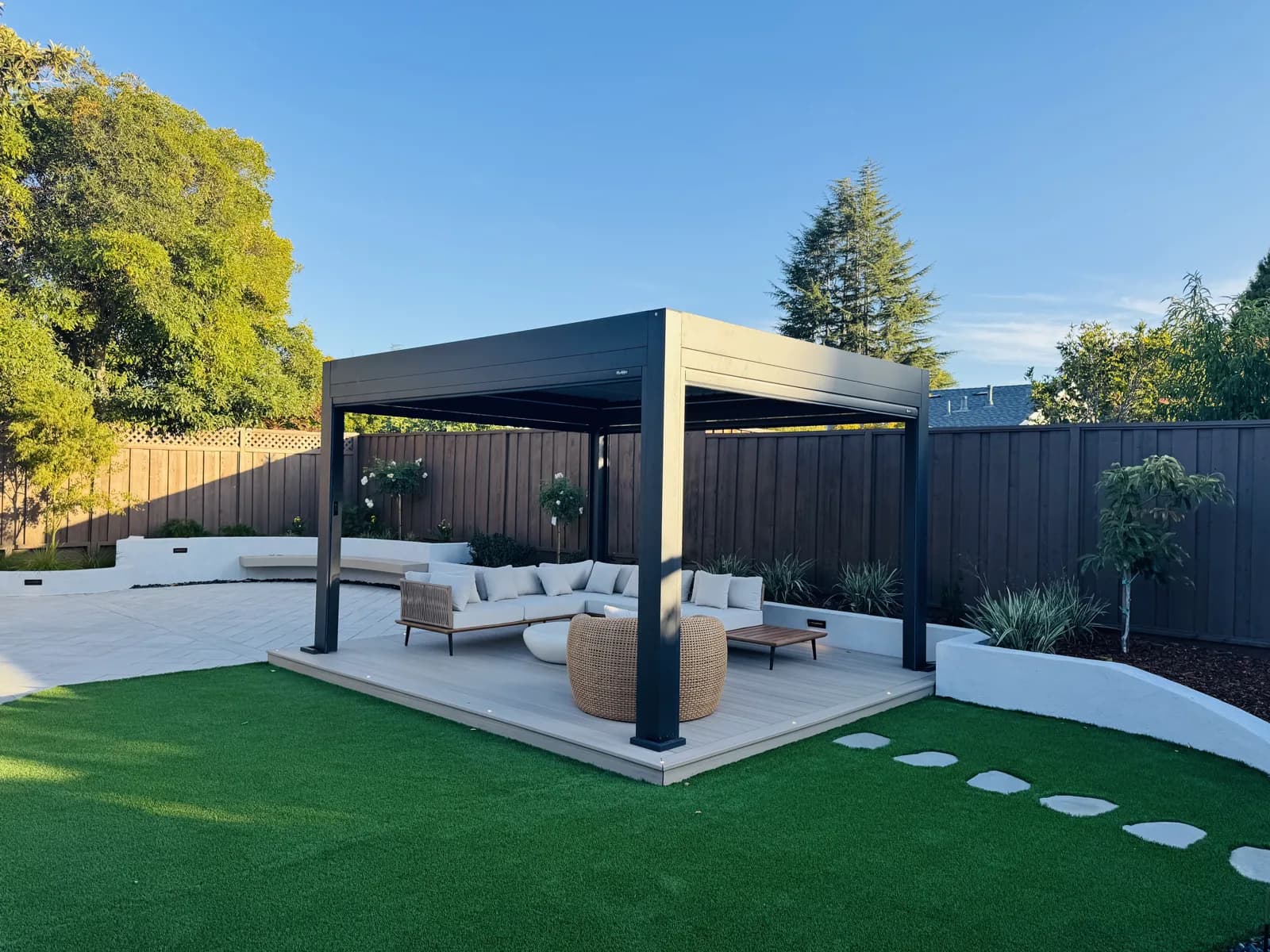 Black louvered pergola on a composite deck with lounge seating, set in an artificial turf lawn